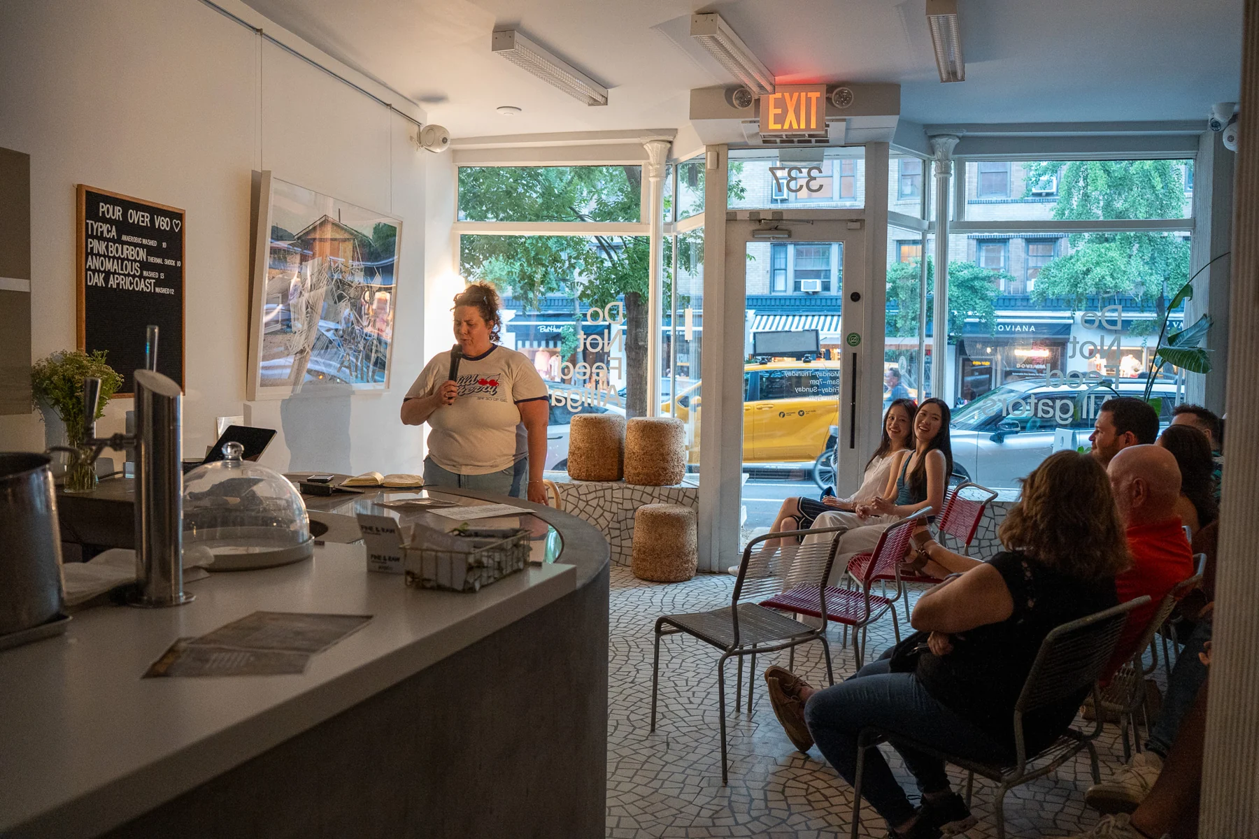 An intimate comedy performance in a bright room, with the audience seated by the windows and a curved counter.