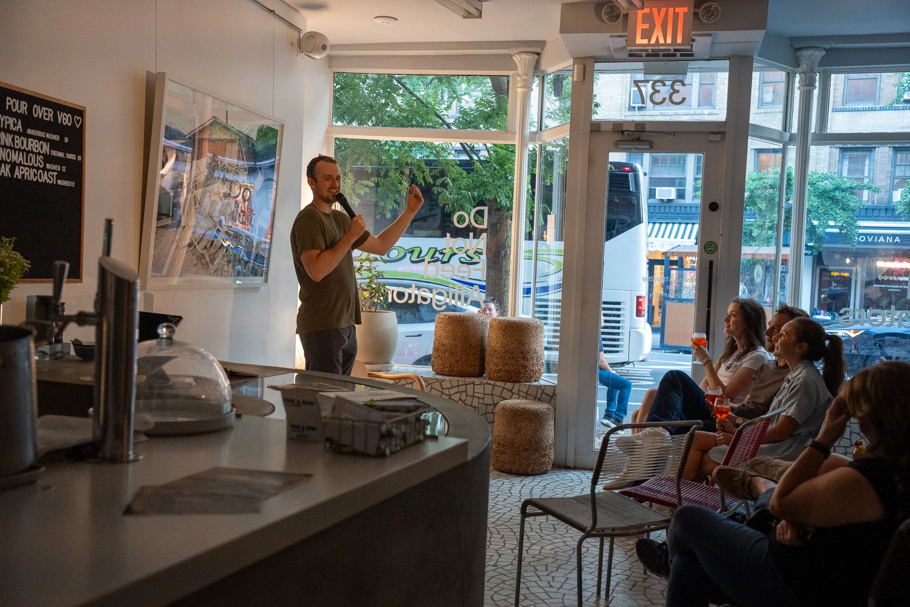 A comedian with a microphone beside large windows, with audience members seated around the room.