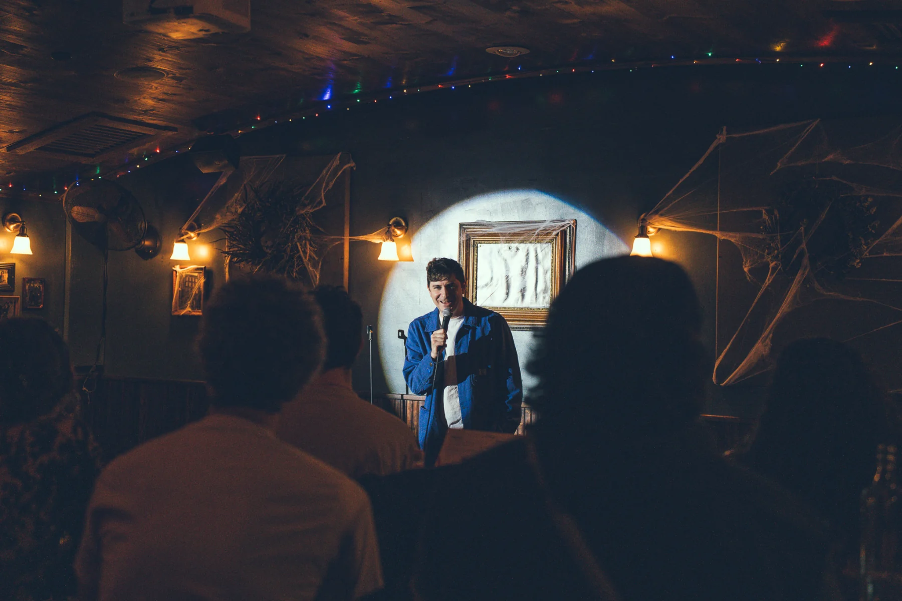 Ryan Beck holding a microphone in a circular spotlight at Comedy T-Rex, with the seated audience in the foreground.