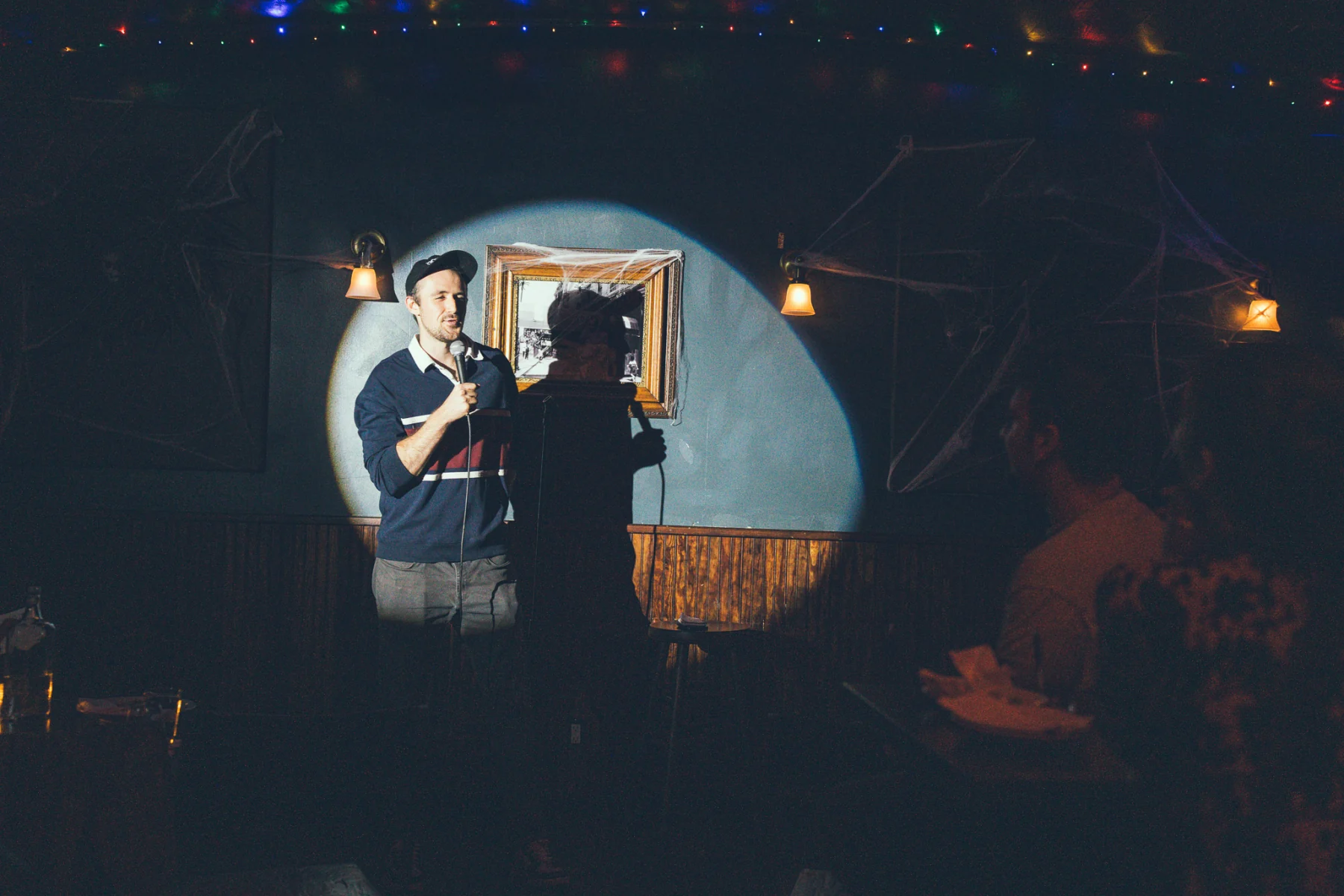Sam Frank Jr. holding a microphone in a circular spotlight at Comedy T-Rex, with the audience in the foreground.