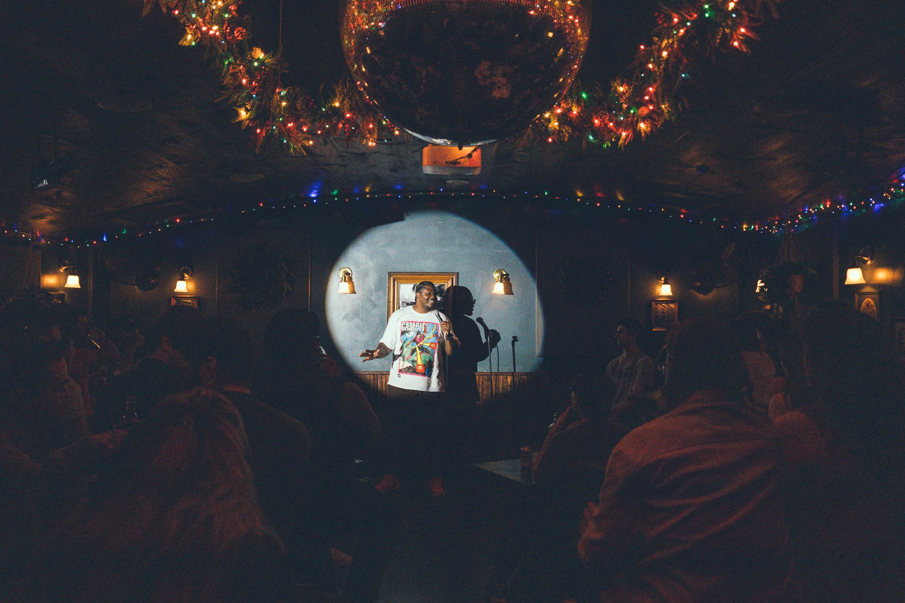 A view through the seated audience toward a spotlighted comedy stage and a chandelier wrapped in lights.